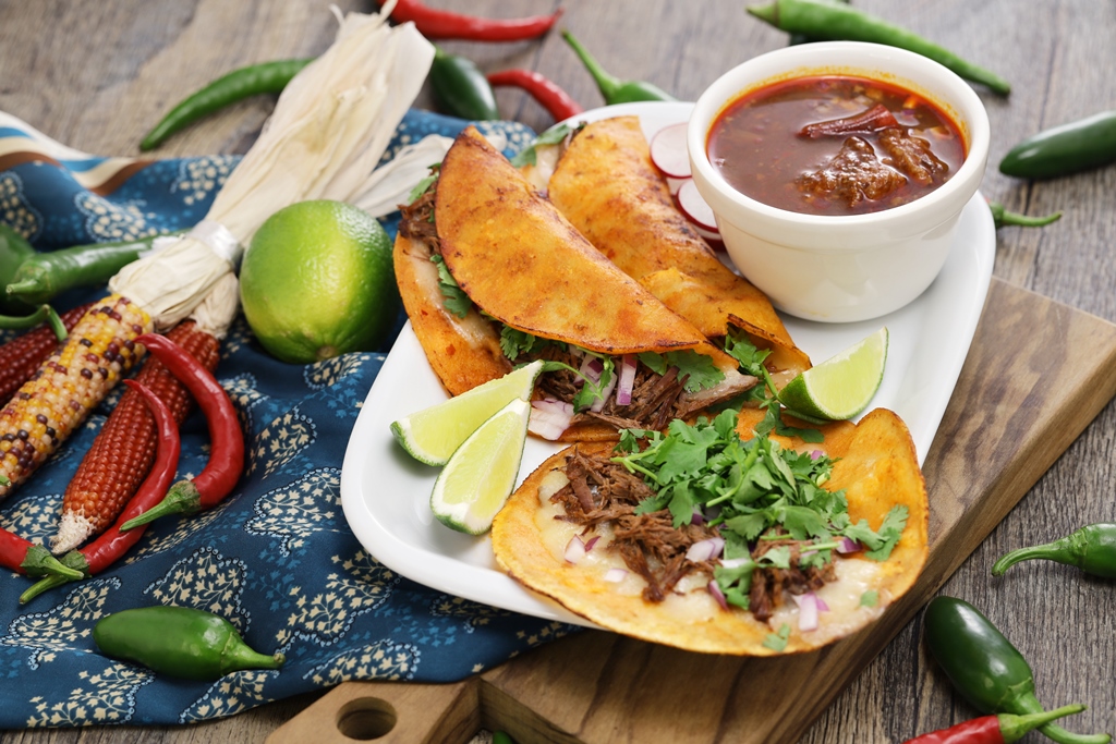 Crispy birria tacos filled with shredded beef, served with consommé, lime wedges, and cilantro on a white plate – Yummy Cooking Treats
