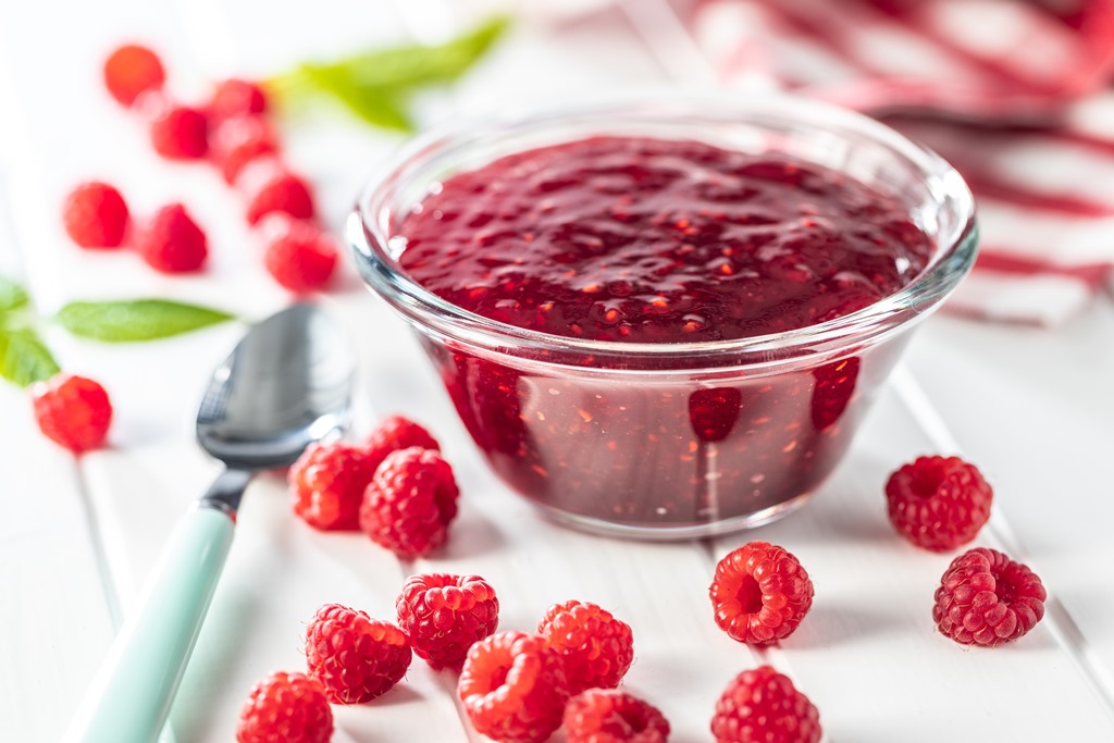 Glass bowl of fresh raspberry sauce surrounded by whole raspberries and a spoon on a white table – Yummy Cooking Treats