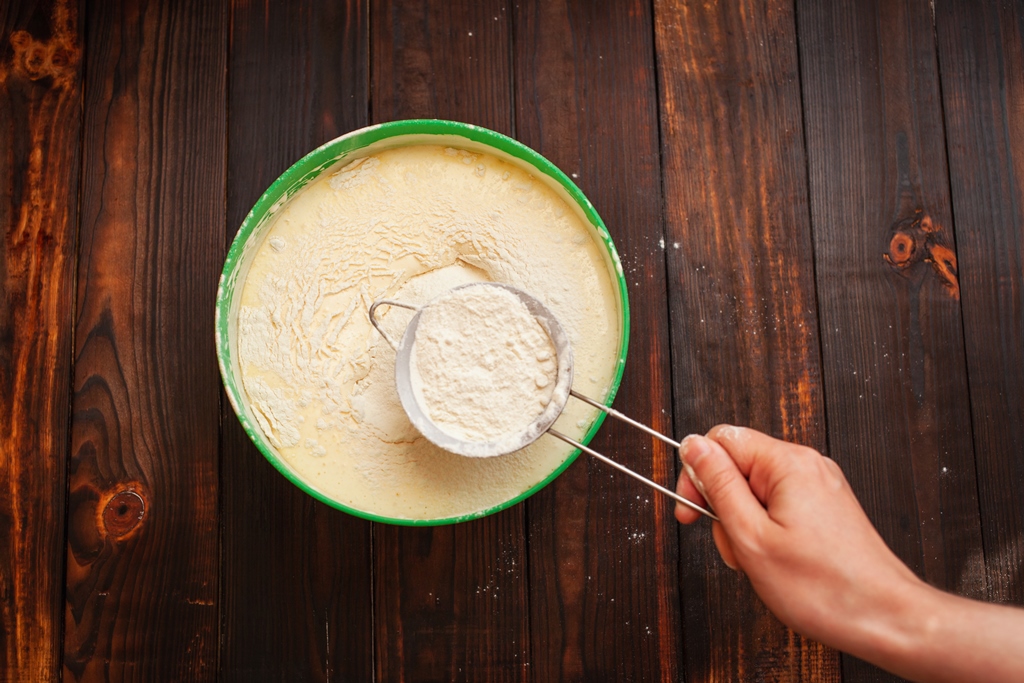 A hand sifting almond flour into pancake batter in a green bowl on a wooden table – Yummy Cooking Treats