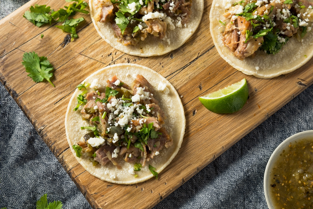 Corn tortillas topped with crispy pork carnitas, chopped cilantro, queso fresco, and lime wedges on a wooden board – Yummy Cooking Treats