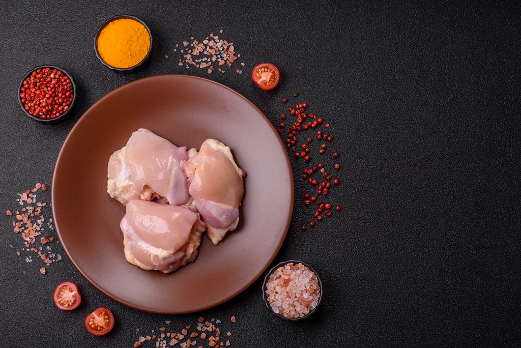 Boneless raw chicken thighs on a brown plate surrounded by spices, salt, and tomatoes on a dark surface – Yummy Cooking Treats