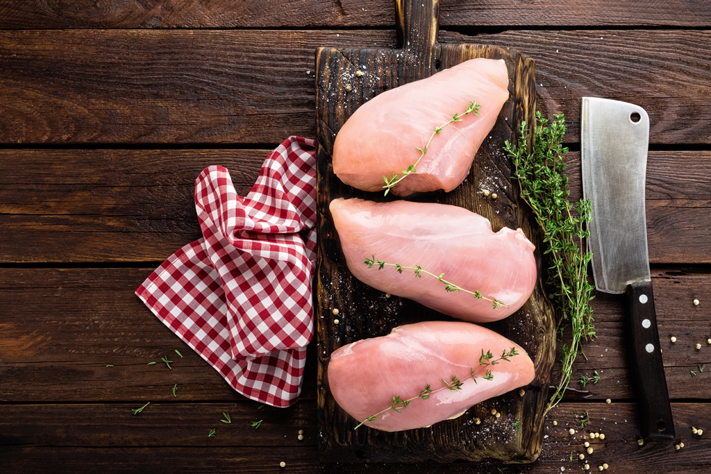 Raw chicken breasts on a wooden board with fresh thyme, a cleaver, and red gingham cloth – prepping for chicken parmesan by Yummy Cooking Treats