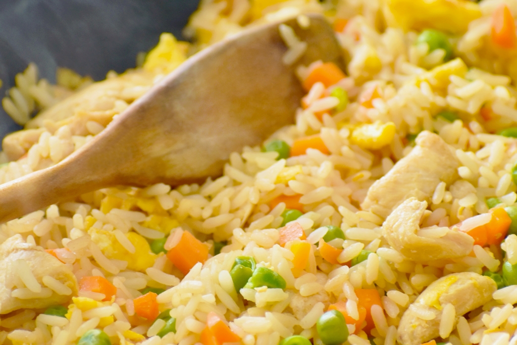 Close-up of egg fried rice with peas, carrots, egg, and tender chicken pieces being stirred with a wooden spoon — Yummy Cooking Treats recipe step