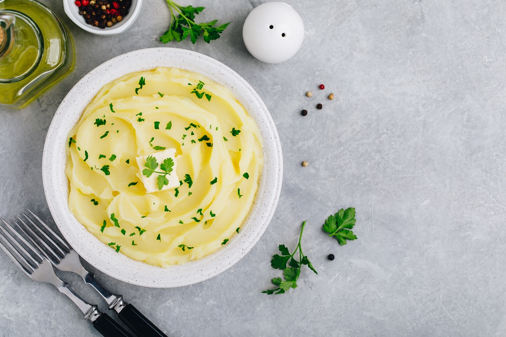 Bowl of creamy mashed potatoes topped with butter and fresh parsley, served with seasonings on a light stone background – Yummy Cooking Treats
