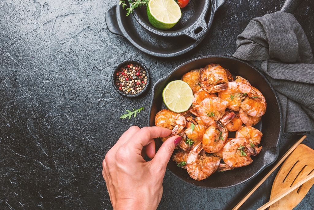 Top view of garlic butter shrimp in a black skillet garnished with lemon and thyme, with a hand picking a piece – Yummy Cooking Treats